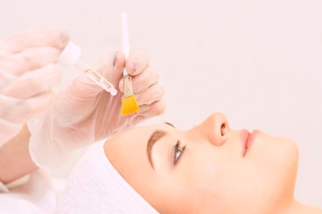 A close-up of a woman lying down with eyes open and hair wrapped in a towel, as a professional wearing gloves prepares to apply a chemical peel using a dropper and a small yellow application brush in a clean spa environment.