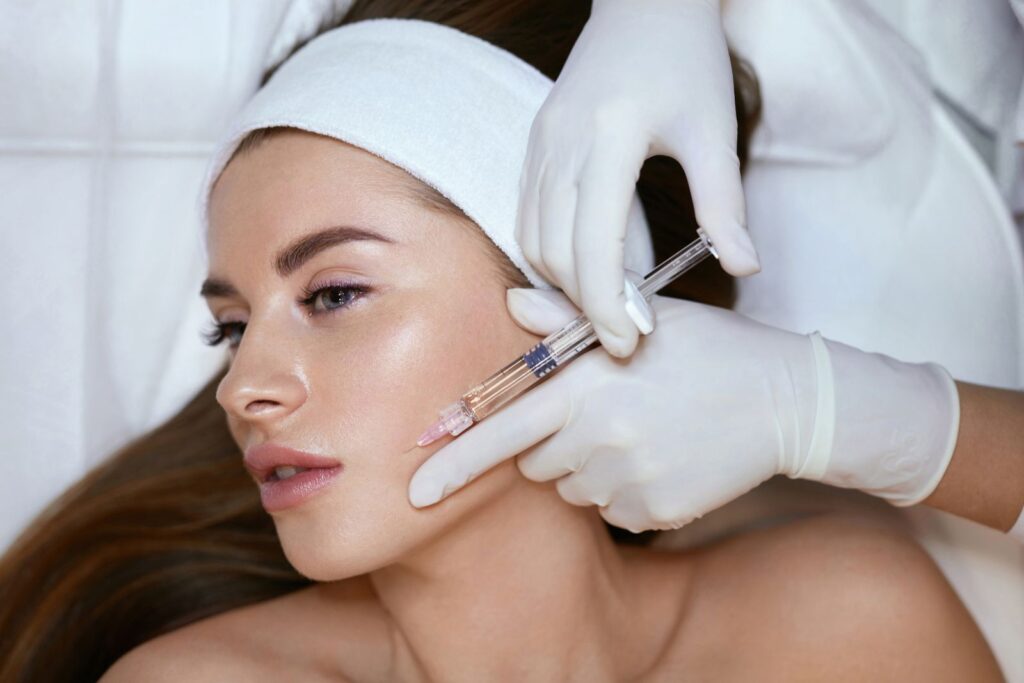 A young woman lies on a treatment bed with a white headband as a medical professional wearing white gloves administers a dermal filler injection to her cheek, part of an anti-aging skincare procedure in a clinical setting.