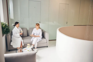 A woman in a white robe sits on a gray sofa speaking with an aesthetic practitioner in uniform inside a modern, minimalistic spa consultation room. They appear relaxed and engaged, with a glass teapot and snacks on a nearby table, set against a clean white interior with numbered treatment room doors in the background.