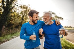 A smiling younger man and older man, both dressed in blue athletic wear, walk arm in arm on a sunlit path while holding water bottles, enjoying a joyful moment together during outdoor exercise.
