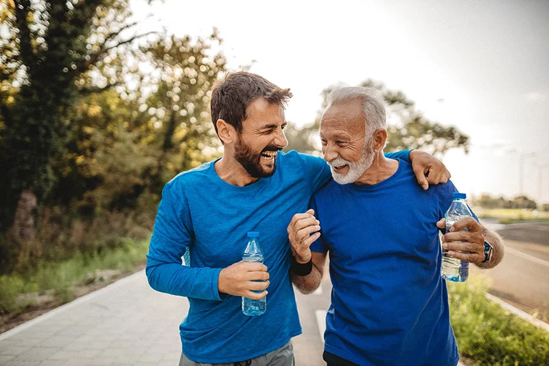 A smiling younger man and older man, both dressed in blue athletic wear, walk arm in arm on a sunlit path while holding water bottles, enjoying a joyful moment together during outdoor exercise.