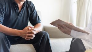 A man sitting on a sofa with hands clasped during a wellness consultation, facing a healthcare provider holding a clipboard, symbolizing a confidential and professional discussion about men's health.