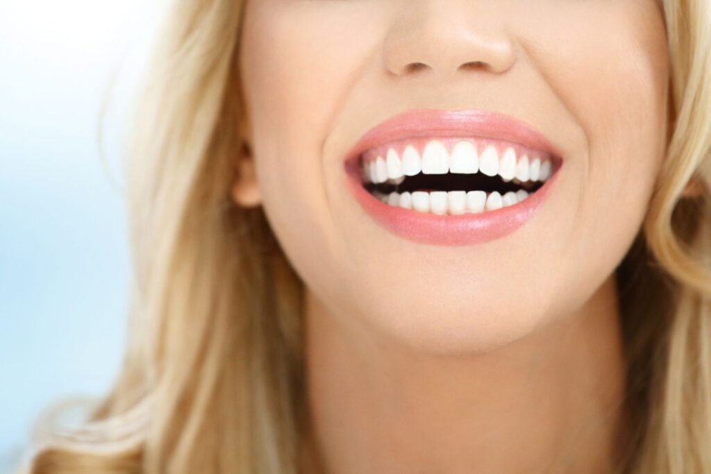 Close-up of a smiling blonde woman showing off her bright white, straight teeth and glossy pink lips, emphasizing dental health, cosmetic dentistry results, or teeth whitening.