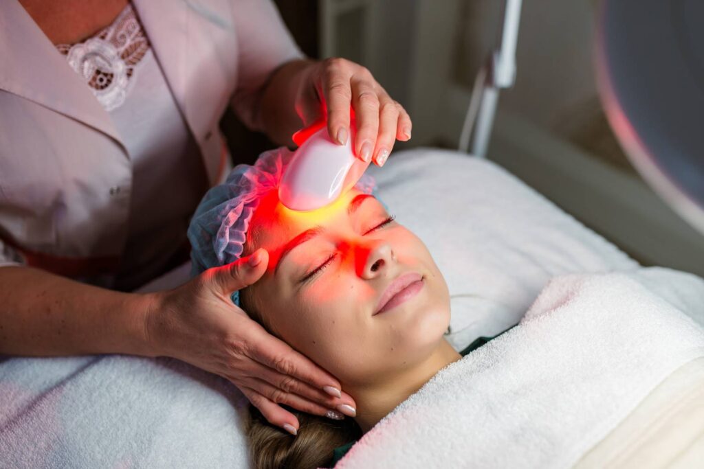 Young woman lying comfortably with eyes closed while receiving a red LED light therapy facial treatment from a professional using a handheld device in a spa or skincare clinic.