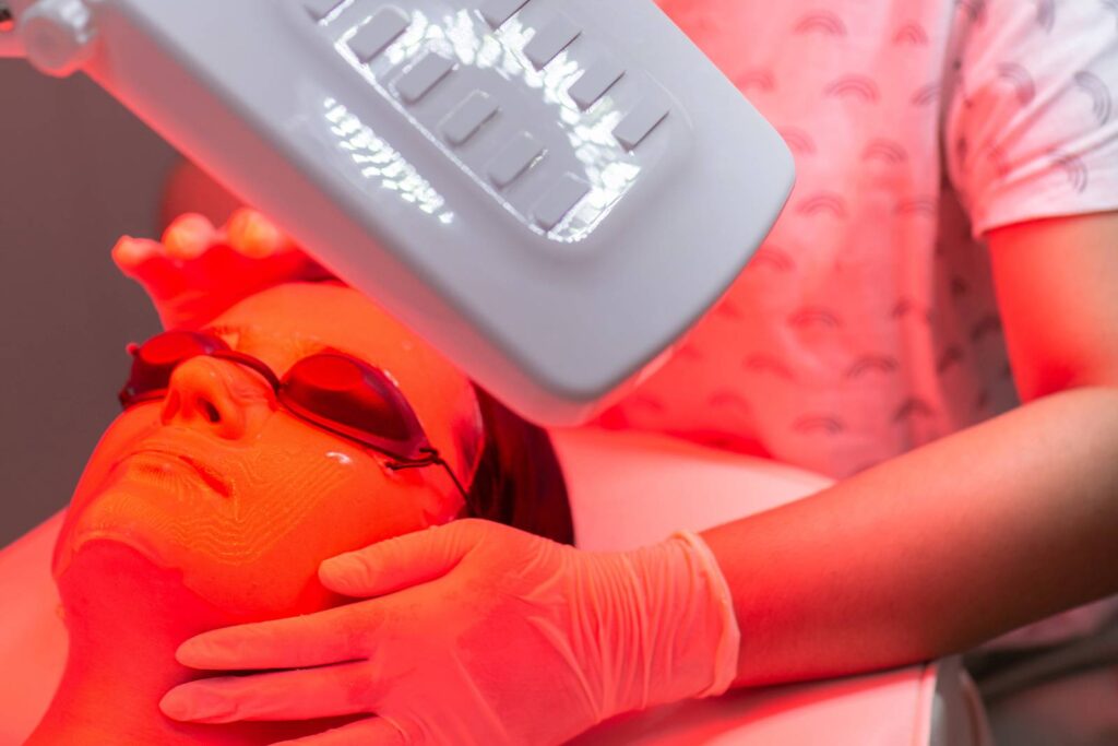 Close-up of a young woman receiving red LED light facial therapy in a beauty salon, lying back with protective goggles and attended by a gloved professional under a glowing red treatment lamp.