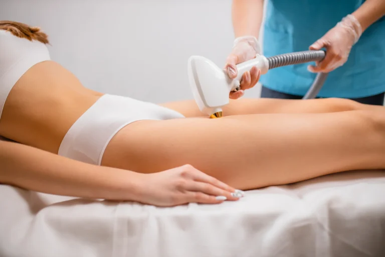 A woman lying on a treatment bed receives a professional Brazilian laser hair removal session. She is wearing white undergarments while a technician in blue scrubs and gloves uses a handheld laser device on her bikini area in a clinical setting.