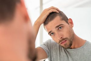 A concerned young man with short hair and a beard looks at his scalp in the mirror while lifting his hair with one hand, inspecting signs of hair thinning or hair loss. He wears a gray t-shirt and appears to be in a bright, indoor setting.