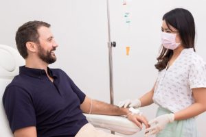 A smiling man sits comfortably in a clinic chair receiving intravenous vitamin therapy through his arm, while a female healthcare worker wearing gloves and a pink face mask monitors the procedure and speaks with him in a calm, sterile environment.
