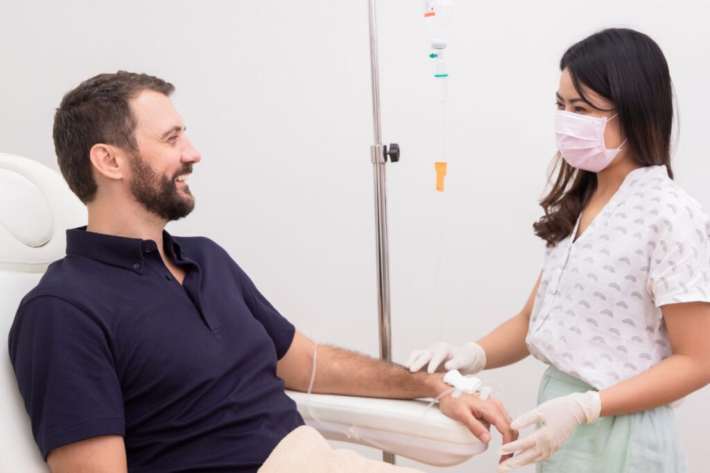 A smiling man sits comfortably in a clinic chair receiving intravenous vitamin therapy through his arm, while a female healthcare worker wearing gloves and a pink face mask monitors the procedure and speaks with him in a calm, sterile environment.